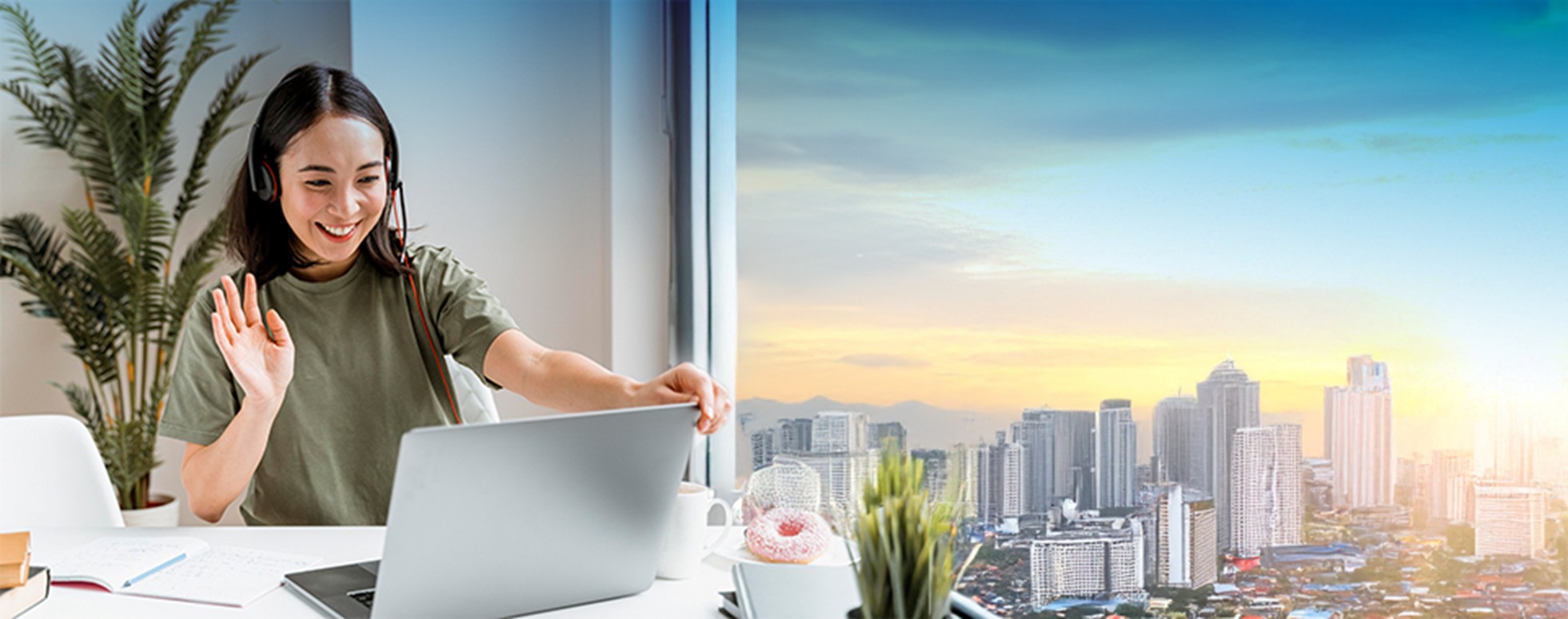 A woman waves during a video call while sitting at a desk with a laptop, notebooks, and a cup of coffee.